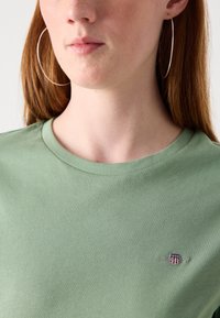 T-shirt in cotone verde con scollo a girocollo, che mostra una texture liscia e un piccolologo ricamato con accenti rossi, bianchi e blu.