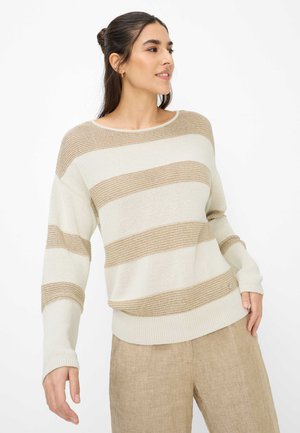 Vrouw draagt beige-wit gestreepte gebreide trui en beige broek, staand met het hoofd naar de zijkant gedraaid tegen een witte achtergrond.