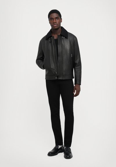 Man in een zwart leren jack met een gespschoen, zwarte broek, witte sokken en zwarte leren instappers, staand tegen een witte achtergrond.