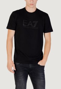 T-shirt nera in cotone con maniche corte, caratterizzata da un logo EA7 testurizzato e la scritta "Emporio Armani" stampata in lettere bold e in contrasto.