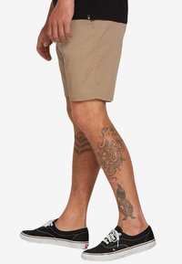 Beige Shorts mit geradem Schnitt, aus leichtem Stoff. Getragen mit schwarzen Sneakers mit weißen Schnürsenkeln und sichtbaren Tattoos am Bein.
