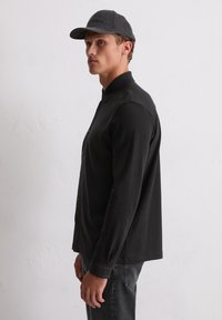 Junger Mann in schwarzem Button-Up-Hemd mit langen Ärmeln und dunkler Jeans, der seitlich an einer weißen strukturierten Wand steht und eine graue Mütze trägt.