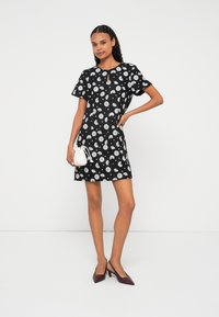 Čierne šaty s kvetinovým vzorom, krátkymi rukávmi, výstrihom s priehlbinou a priliehavým strihom. Modelka drží malú bielu kabelku a má na sebe vínové lodičky.