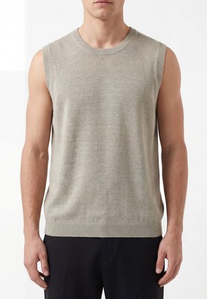 Maglione senza maniche in maglia beige chiaro indossato da un uomo in piedi con le braccia rilassate, abbinato a pantaloni neri su sfondo uniforme.