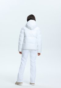 Doudoune blanche avec capuche, présentant des sections matelassées et une coupe courte. Portée avec un pantalon blanc et des bottes beiges, sur un fond uni.