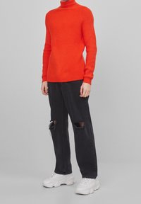 Jersey de cuello alto acanalado rojo con mangas largas, combinado con jeans negros desgastados y zapatillas blancas gruesas, contra un fondo neutro.