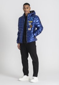 Blå puffjacka med en glänsande finish, med flera märken, dragkedja fram och huva. Svarta byxor och vita sneakers fulländar looken.