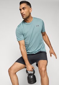 Camiseta atlética de manga corta color azul claro con un patrón diagonal texturizado, combinada con pantalones cortos negros. Persona sosteniendo un kettlebell negro.