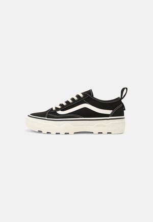 Czarny niski sneaker z białym falistym paskiem, białymi sznurowadłami, grubą białą podeszwą i pętelką na pięcie, na jasnoszarym tle.