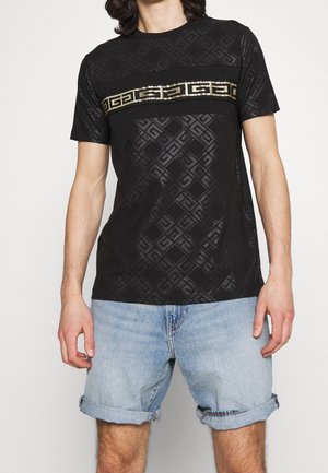 Hombre con camiseta negra estampada con una franja geométrica dorada en el pecho y pantalones cortos de mezclilla azul claro arremangados.
