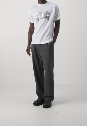 Mann trägt weißes, kurzärmeliges T-Shirt mit dem Schriftzug "ÉTUDES", dunkelgraue weite Hose und schwarze Schuhe, steht vor einem einfarbigen Hintergrund.