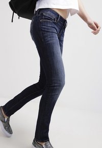 Jeans skinny azul oscuro con cinco bolsillos, áreas desgastadas y detalles de costura sutiles, combinados con zapatos metálicos de tipo slip-on.