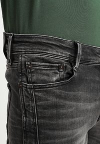 Vaqueros de mezclilla negros con un corte ajustado, textura descolorida, cinco bolsillos y detalles de herrajes en tono cobre. Llevados con una blusa verde.