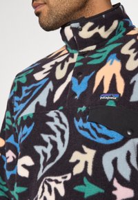 Sudadera de forro polar con cuello alto. El diseño presenta hojas en colores pastel sobre un fondo oscuro. Bolsillo en el pecho negro con logo.