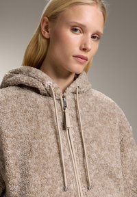 Beżowa kurtka z sztucznego futra z kapturem, z suwakiem z przodu i z zapięciem na troczki, o teksturowanej tkaninie i luźnym kroju. Gładkie troczki i metalowe akcenty.