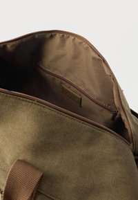 Intérieur de sac en toile vert olive avec doublure en tissu marron, comprenant une poche zippée et une étiquette en cuir. Détails de couture robustes visibles.