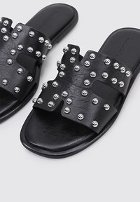 Sandalias deslizantes de cuero negro con una amplia correa superior adornada con tachuelas metálicas plateadas. Suela plana con una textura suave y diseño abierto.