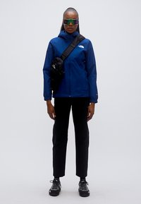 Μπλε αδιάβροχο μπουφάν με κουκούλα, μαύρο σακίδιο, μαύρο παντελόνι και μαύρα παπούτσια πεζοπορίας. Το μοντέλο φοράει ανακλαστικά γυαλιά ηλίου.