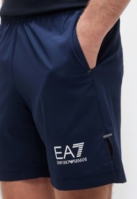 Άνδρας που φοράει ναυτικό μπλε σορτς Emporio Armani EA7 με χέρι στην δεξιά τσέπη, σε λευκό φόντο.