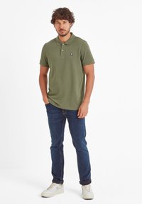 Man stands facing forward wearing olive green polo shirt, dark blue jeans, white sneakers, with one hand in pocket against white background.