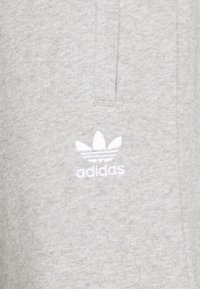 Svetle šedá mikina vyrobena z měkké tkaniny. Na levé straně hrudníku se nachází bílé vyšívané logo Adidas s trojlístkem.