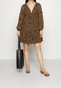 Vestido de manga larga con escote en V profundo en estampado de paisley con tonos terrosos, con volantes en capas en el dobladillo, combinado con un bolso negro y sandalias transparentes.