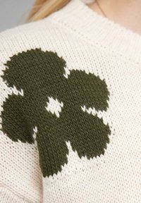 Knitted sweater in cream with a large dark green floral pattern on the shoulder. Features a textured, soft material and ribbed neckline.