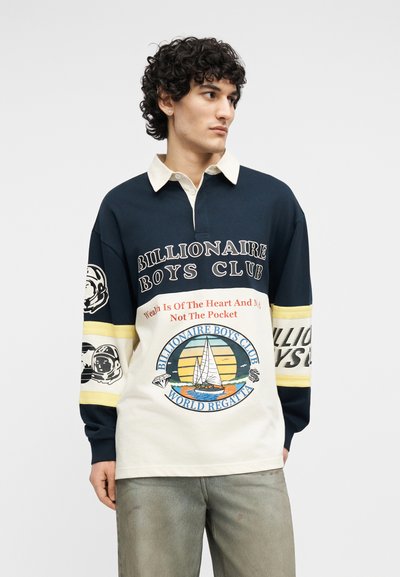 Άνδρας με σγουρά μαλλιά που φοράει μπλε-ναυτικό και κρεμ πουκάμισο με μακριά μανίκια, με κείμενο "Billionaire Boys Club" και εικόνα ιστιοφόρου.