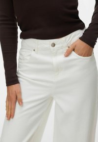 Personne portant un jean blanc taille haute avec des poches avant, une main dans la poche, et un haut à manches longues marron foncé visible de la taille au milieu de la cuisse.