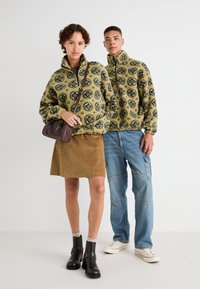 Puloveri din fleece verzi cu modele florale bleumarin, asortați cu o fustă din catifea groasă maro și blugi cargo albaștri. Se poartă cizme negre și adidași.