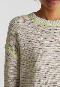 Stickad tröja i krämfärg med flerfärgade fläckar, med rund hals och ljusgröna accenter längs halsringningen och ärmarna.