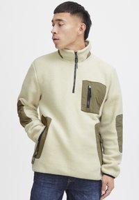 Fleece pullover in off-white with olive green accents. Features a half-zip neck, chest pocket, and elbow patches. Soft texture.