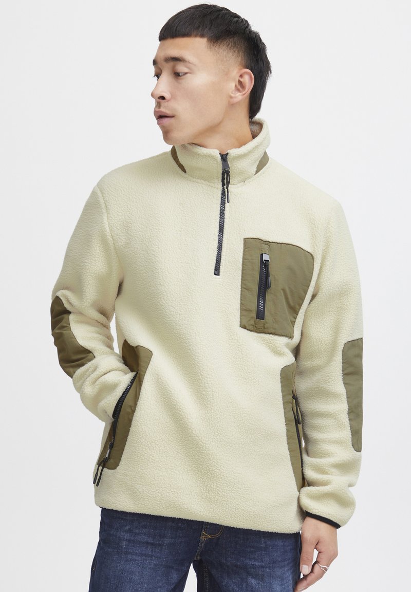Fleece pullover in off-white with olive green accents. Features a half-zip neck, chest pocket, and elbow patches. Soft texture.