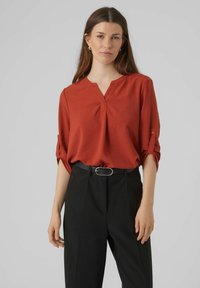 Blusa rossa con scollatura a V e maniche arrotolate. Indossata con pantaloni neri a vita alta e una cintura nera con una fibbia circolare.