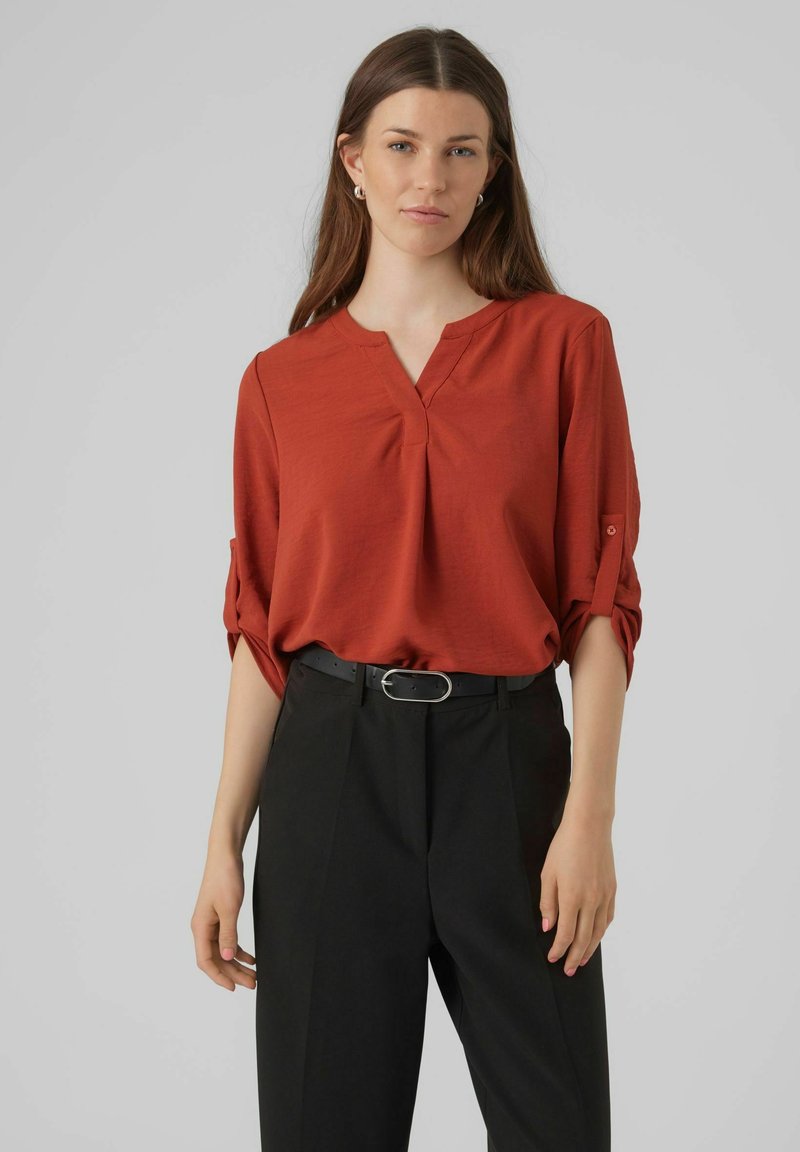 Rode blouse met een V-hals en opgerolde mouwen. Ggedragen met zwarte high-waisted trousers en een zwarte riem met een ronde gesp.