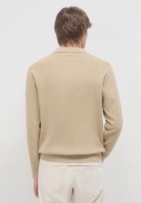 Mann mit kurzen braunen Haaren, der einen beigen langärmeligen Pullover und off-white Hosen trägt, mit dem Gesicht von einer einfachen hellen Hintergrund abgewandt.