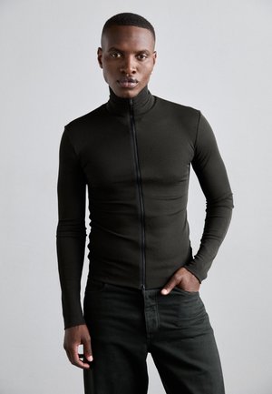 Suéter ajustado de manga larga en tela verde oscuro con cuello alto y cremallera frontal, combinado con pantalones verdes oscuros. Textura suave.