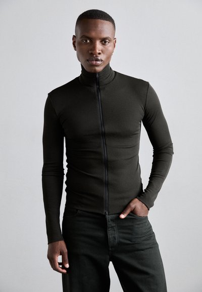 Pull à manches longues ajusté en tissu vert foncé avec un col haut et une fermeture éclair à l'avant, associé à un pantalon vert foncé. Texture lisse.