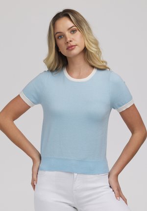 Młoda kobieta o blond włosach ma na sobie jasnoniebieską bluzkę z krótkim rękawem z białym wykończeniem i białe spodnie, stoi ze złożonymi rękami na biodrach.