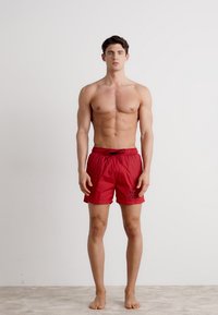 Shorts de bain rouges de couleur unie, avec une taille à cordon de serrage et un logo sur le devant. Le matériau semble léger et à séchage rapide.