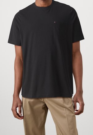 Mann trägt schwarzes Kurzarm-T-Shirt mit Brusttasche und beige Hose vor weißem Hintergrund.