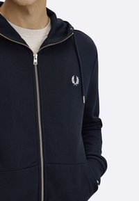 Granatowa bluza z kapturem zapinana na zamek, wykonana z miękkiego materiału. Posiada srebrny zamek błyskawiczny, logo z przodu oraz dwie boczne kieszenie. Kaptur z regulowanymi sznurkami.