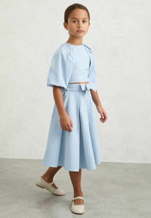 Junges Mädchen in hellblauem bauchfreiem Oberteil mit Puffärmeln und passendem Midi-Rock mit Schleife, trägt beige Mary Jane Schuhe, steht auf grauem Boden.