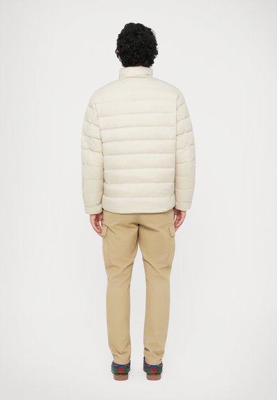 Giacca crema imbottita con design a righe orizzontali, colletto alto e polsini elasticizzati, abbinata a pantaloni cargo beige e scarpe multicolori.