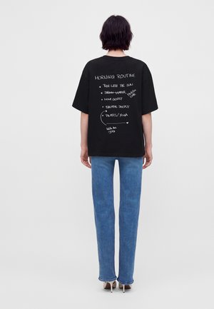 Άτομο με κοντά σκούρα μαλλιά φοράει μαύρο T-shirt με λευκή χειρόγραφη επιγραφή "ρουτίνα πρωινού" και μπλε τζιν, στέκεται μπροστά σε ένα απλό φόντο.