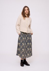 Donna che indossa un cardigan beige con bottoni, una gonna midi floreale blu scuro, calze nere e scarpe oxford marrone scuro, in piedi con le mani nelle tasche.