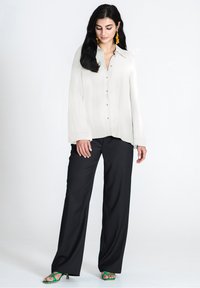 Blusa grigia chiara con chiusura a bottoni, maniche lunghe e vestibilità ampia, abbinata a pantaloni neri a palazzo e sandali con tacco verdi.