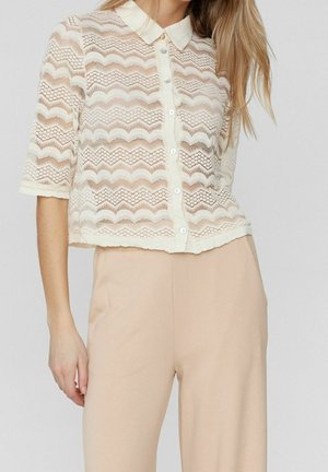 Frau trägt eine cremefarbene Spitzenbluse mit kurzen Ärmeln und Knöpfen sowie beige hoch taillierte Hosen vor einem einfarbigen Hintergrund.