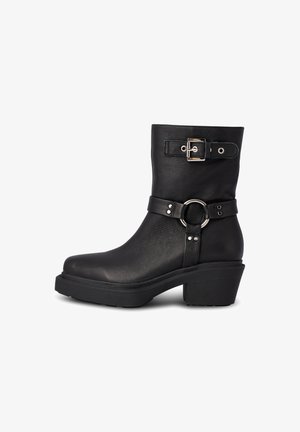 Bottines en cuir noir avec un talon épais, accents de boucle, détail en forme d'anneau circulaire sur la sangle, surface subtilement texturée et semelle en caoutchouc.