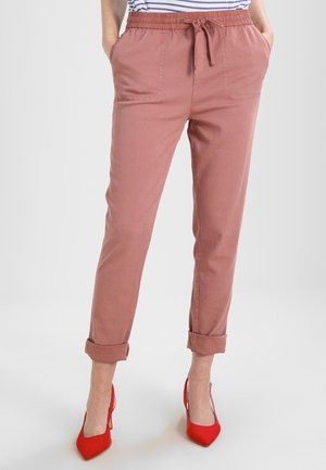 Vrouw draagt stoffige roze broek met omgeslagen pijpen, trekkoord in de taille en voorzakken, gecombineerd met felrode hoge hakken.